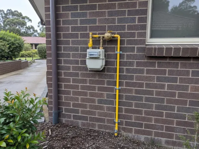 Yellow gas meter and pipework mounted on a brick house wall beside a window in Pennant Hills